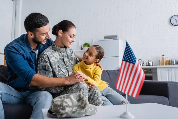 Smiling family sitting near mother in military uniform and blurred american flag at home — Stock Photo