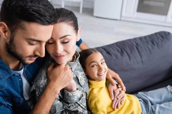 Man with closed eyes holding hand of wife in military uniform near blurred child at home — Stock Photo