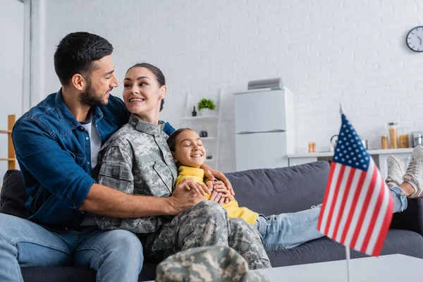 Positiver Mann umarmt Ehefrau in Tarnuniform neben Tochter und verschwommener amerikanischer Flagge zu Hause — Stockfoto