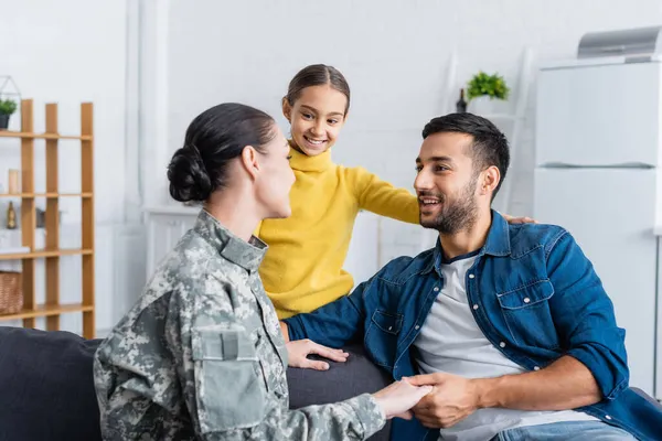 Frau in Militäruniform hält Hand ihres Mannes neben lächelnder Tochter zu Hause — Stockfoto