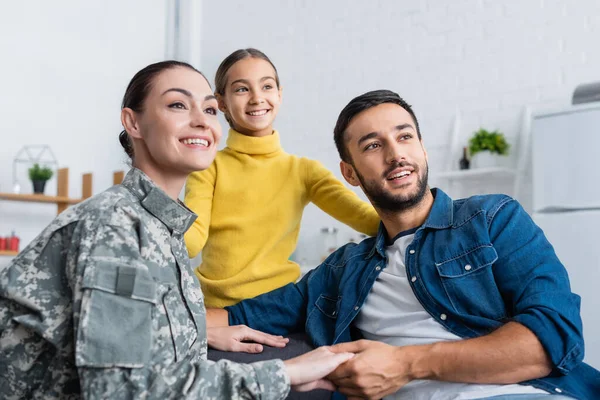 Mujer positiva en uniforme militar cogida de la mano del marido cerca del niño mirando hacia otro lado en casa - foto de stock