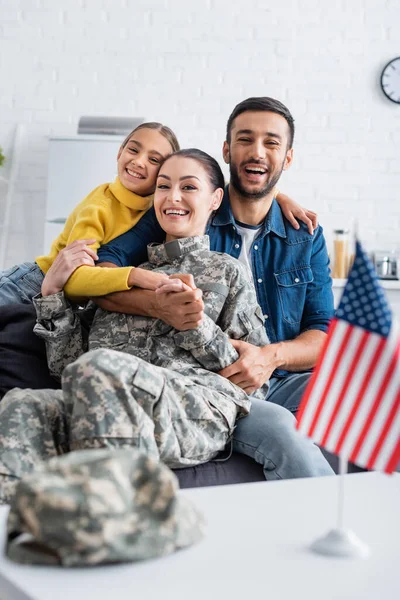 Glückliche Familie umarmt Mama in Militäruniform und schaut in die Kamera neben der amerikanischen Flagge zu Hause — Stockfoto