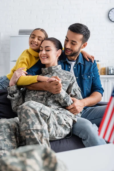 Ragazzo sorridente che abbraccia la madre in uniforme militare vicino al padre e sfocata bandiera americana a casa — Foto stock