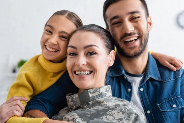 Familia positiva mirando a la cámara cerca de la madre en uniforme militar en casa - foto de stock