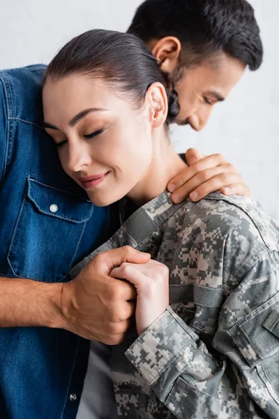 Blurred man hugging and holding hand of wife in military uniform at home — Stock Photo