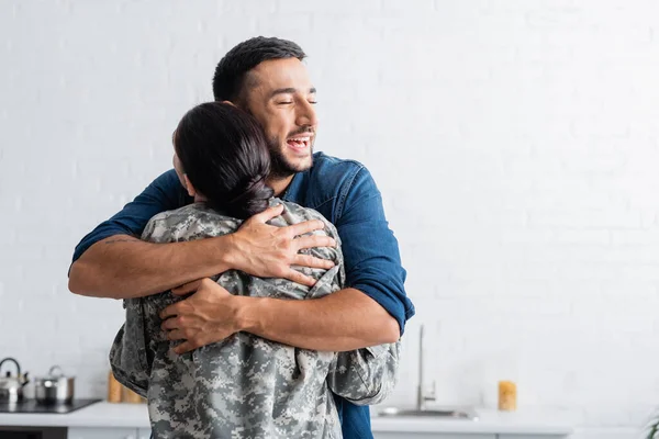 Feliz hombre abrazando esposa en uniforme militar en la cocina en casa - foto de stock