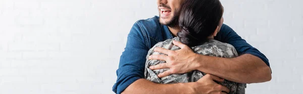 Vista recortada del hombre alegre abrazando esposa en uniforme militar en casa, pancarta - foto de stock
