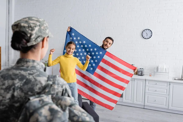 Uomo felice che tiene la bandiera americana vicino al bambino mostrando sì gesto e madre sfocata in uniforme militare a casa — Foto stock