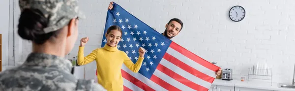 Familia feliz con bandera americana mirando a la mujer en uniforme militar en casa, pancarta - foto de stock