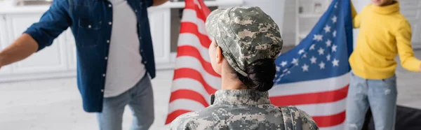 Mujer en uniforme militar de pie cerca de la familia borrosa con bandera americana en casa, pancarta — Stock Photo