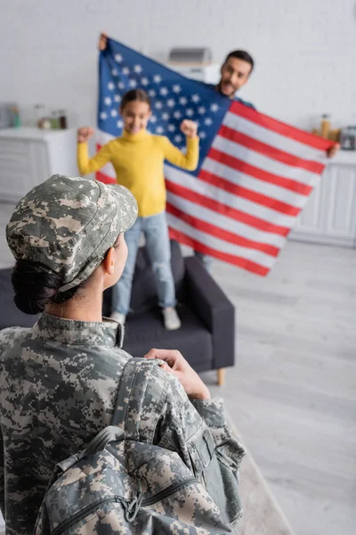 Mulher de uniforme militar com mochila perto da família borrada segurando bandeira americana na cozinha — Fotografia de Stock