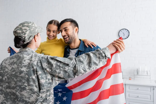 Hombre y niño sosteniendo bandera americana cerca de la mujer en uniforme militar en casa - foto de stock