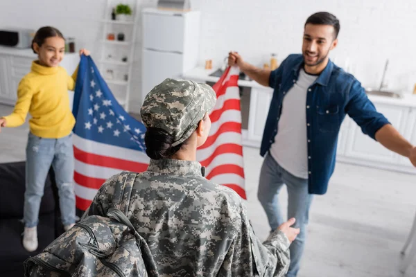 Donna in uniforme militare in piedi vicino a marito e bambino sfocati con bandiera americana a casa — Foto stock