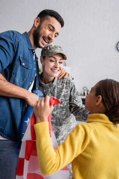 Lächelnder Mann umarmt Ehefrau in Militäruniform neben Mädchen mit amerikanischer Flagge zu Hause — Stockfoto