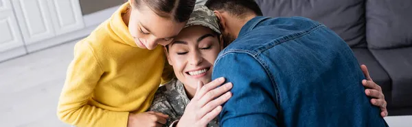 Mujer feliz en uniforme militar abrazando marido cerca de la hija en casa, bandera - foto de stock