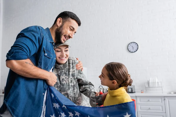 Мужчина обнимает жену в военной форме рядом с дочерью в американском флаге на кухне — стоковое фото