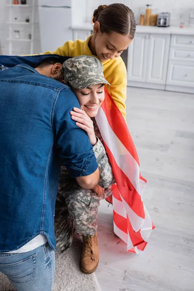 Mann umarmt Ehefrau in Militäruniform neben Tochter mit amerikanischer Flagge zu Hause — Stockfoto