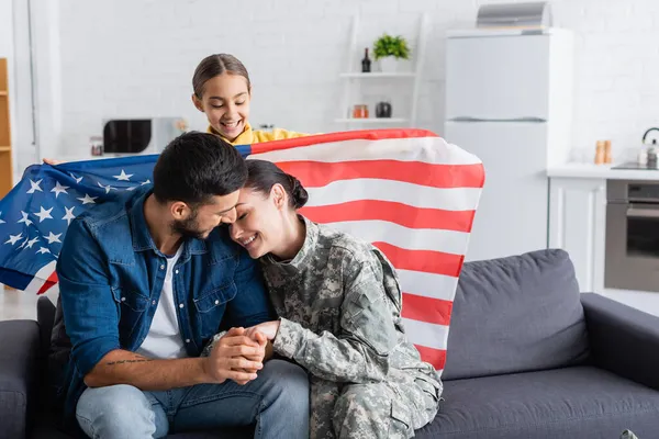 Sorrindo menina pré-adolescente segurando bandeira americana perto de mãe em uniforme militar e pai no sofá — Fotografia de Stock