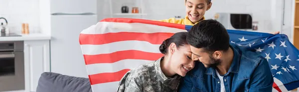 Hija preadolescente sosteniendo bandera americana cerca de mamá en uniforme militar y papá en casa, pancarta - foto de stock