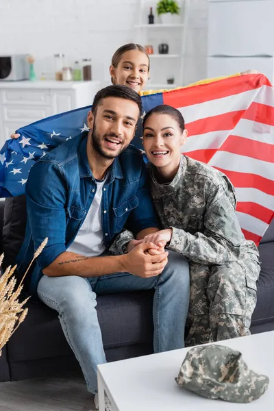 Donna felice in uniforme militare che tiene per mano il marito vicino bambino sorridente con bandiera americana guardando la fotocamera a casa — Foto stock