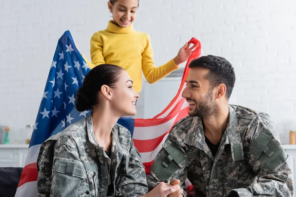 Веселые родители в военной форме смотрят друг на друга рядом с дочерью с американским флагом дома — стоковое фото