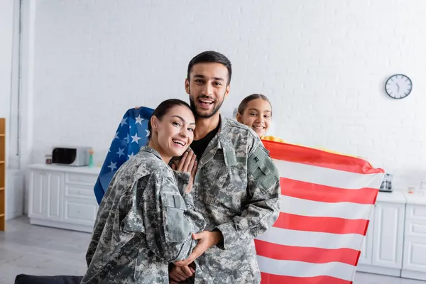 Glücklicher Mann und Frau in Militäruniform blickt in die Kamera neben Tochter mit amerikanischer Flagge zu Hause — Stockfoto