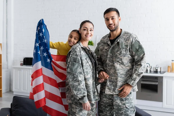 Padres en uniforme militar tomados de la mano cerca de un niño preadolescente con bandera americana en casa - foto de stock