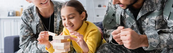 Усміхнена дитина грає в дерев'яні блоки біля батьків у військовій формі вдома, банер — стокове фото