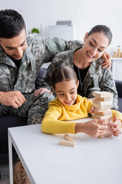 Homem de uniforme militar abraçando esposa perto de criança jogando blocos de madeira jogo em casa — Fotografia de Stock