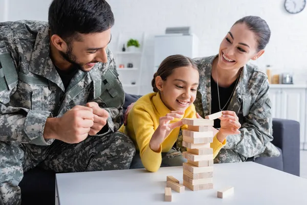 Des parents heureux en uniforme militaire montrant un geste oui près d'un enfant jouant au jeu des blocs de bois à la maison — Photo de stock