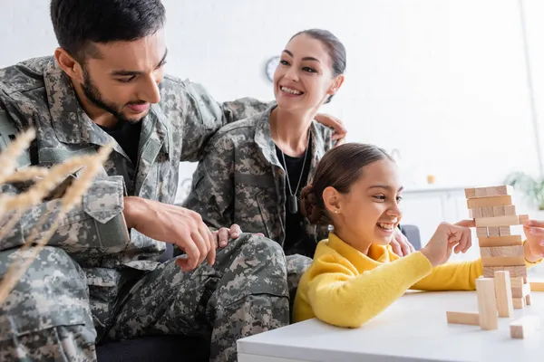 Smiling kid playing wood blocks game near parents in military uniform at home — Stock Photo