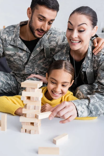 Cheerful woman in military uniform pointing at wood blocks game near husband and kid at home — Stock Photo