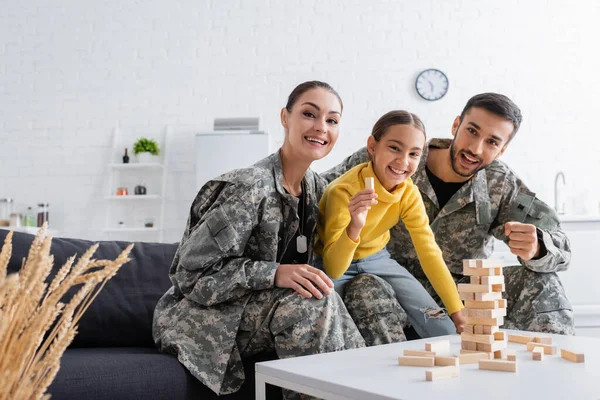 Smiling parents in camouflage uniform sitting near child with wood block near game at home — Stock Photo