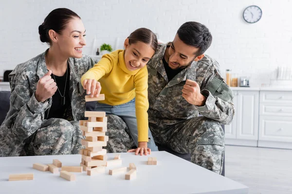 Genitori sorridenti in uniforme militare che mostrano sì gesto vicino bambino giocare blocchi di legno gioco a casa — Foto stock