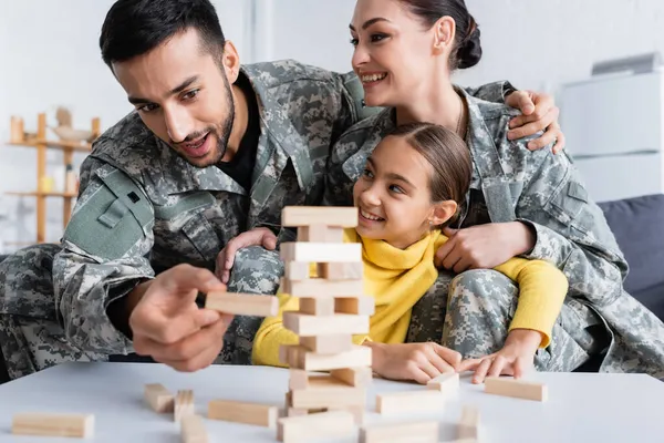 Hombre en uniforme militar jugando bloques de madera borrosa juego cerca de la esposa y el niño en casa - foto de stock