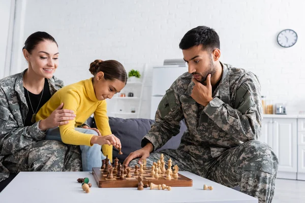 Pensivo pai em camuflagem uniforme jogando xadrez perto sorrindo mãe e filho em casa — Fotografia de Stock