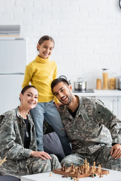 Criança feliz de pé perto dos pais em uniforme militar olhando para a câmera perto de xadrez em casa — Fotografia de Stock