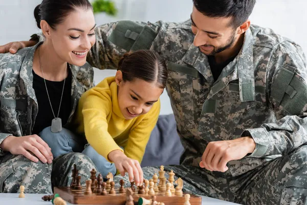 Criança sorrindo jogando xadrez perto dos pais em uniforme de camuflagem em casa — Fotografia de Stock