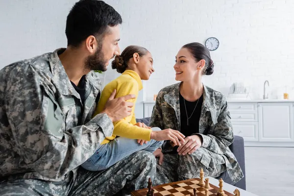 Positive parents in military uniform sitting near daughter and chess at home — Stock Photo