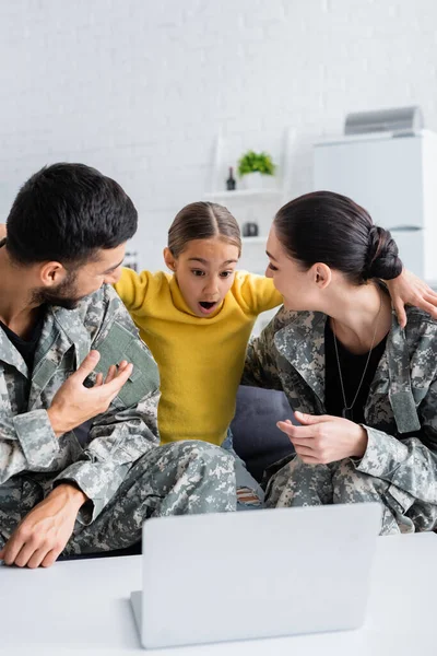 Amazed kid looking at blurred laptop while hugging parents in military uniform at home — Stock Photo