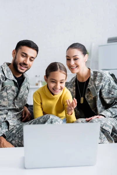 Lächelndes Kind mit verschwommenem Laptop neben positiven Eltern in Militäruniform zu Hause — Stockfoto