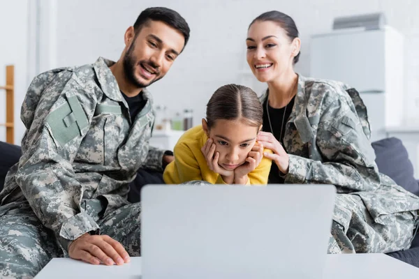 Дитина дивиться на ноутбук біля розмитих батьків у військовій формі на дивані — стокове фото