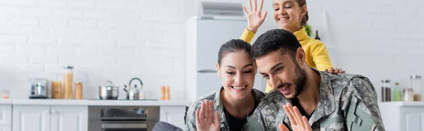 Sonrientes padres en uniforme militar agitando las manos cerca de un niño preadolescente en casa, pancarta - foto de stock