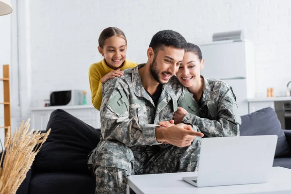 Hombre sonriente en uniforme de camuflaje apuntando a la computadora portátil cerca de la esposa y el niño preadolescente en casa - foto de stock