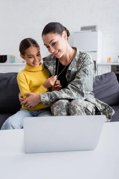 Mère souriante en camouflage étreignant fille préadolescente avec des étiquettes de chien près d'un ordinateur portable sur la table à la maison — Photo de stock