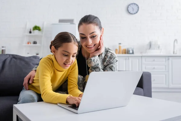 Lächelndes Mädchen mit Laptop neben Mutter in Militäruniform zu Hause — Stockfoto