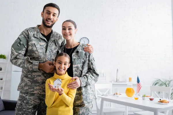 Smiling parents in military uniform standing near daughter with model of house in kitchen — Stock Photo