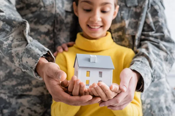 Model of house on hands of blurred kid near parents in military uniform at home — Stock Photo