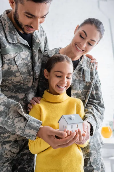 Smiling parents in military uniform holding model of house near child at home — Stock Photo