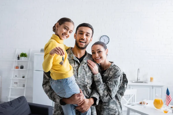 Alegre padres en uniforme militar sosteniendo hija con llave en casa - foto de stock
